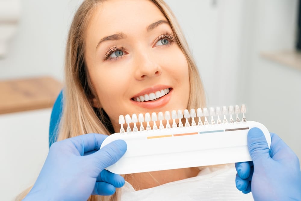 A dentist helps a smiling patient choose a shade for her teeth using a color guide while she looks into a handheld mirror - Dentist Atlanta