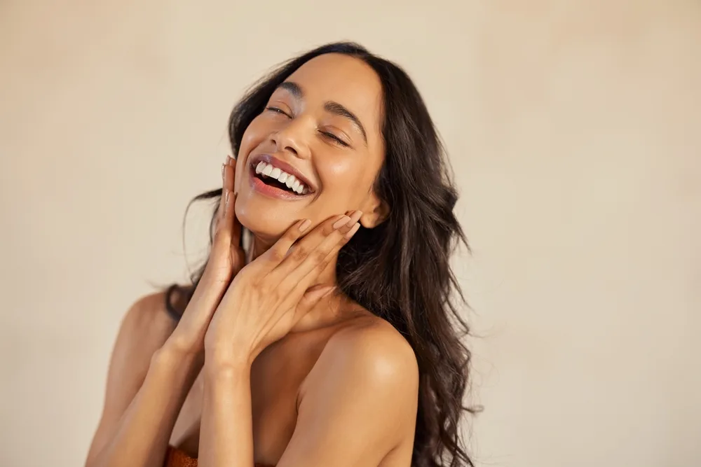 Radiant Woman With a Beautiful, Natural Smile – Cosmetic Dentistry Buckhead GA A joyful woman laughs with her hands gently touching her face, showcasing a smooth, glowing smile that highlights natural beauty – Cosmetic Dentistry Buckhead GA.