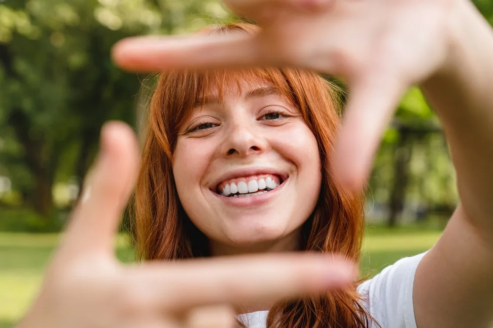 Young Woman Highlighting Her Fresh, Beautiful Smile – Cosmetic Dentistry Buckhead GA A smiling woman frames her face with her hands outdoors, showing off a bright, natural smile that reflects confidence and joy – Cosmetic Dentistry Buckhead GA.