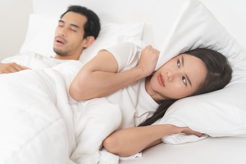 A woman looks frustrated in bed, covering her ears with a pillow while a man beside her snores loudly - Dentist Atlanta A woman looks frustrated in bed, covering her ears with a pillow while a man beside her snores loudly - Dentist Atlanta