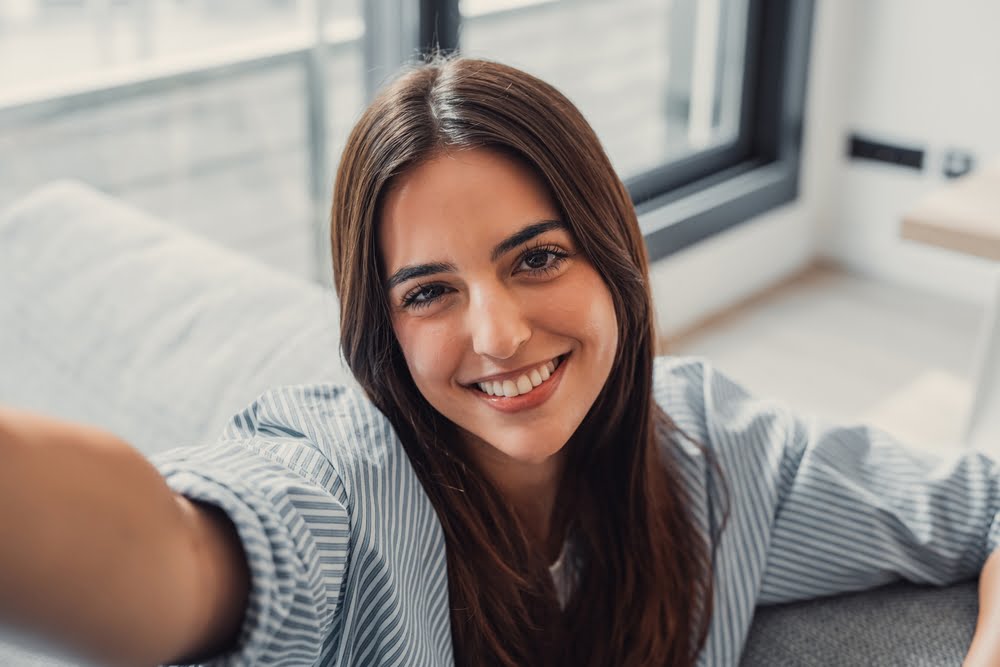 A woman with long hair smiles brightly while taking a selfie indoors - Dentist Atlanta A woman with long hair smiles brightly while taking a selfie indoors - Dentist Atlanta