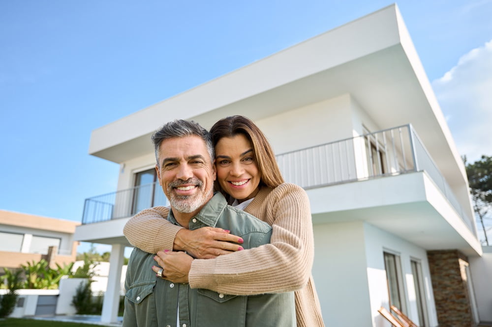 A happy couple smiling outside their modern home, with the woman hugging the man from behind - Dentist Atlanta A happy couple smiling outside their modern home, with the woman hugging the man from behind - Dentist Atlanta
