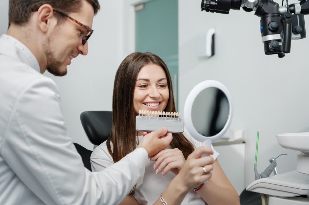 A dentist helps a smiling patient choose a shade for her teeth using a color guide while she looks into a handheld mirror - Dentist Atlanta A dentist helps a smiling patient choose a shade for her teeth using a color guide while she looks into a handheld mirror - Dentist Atlanta