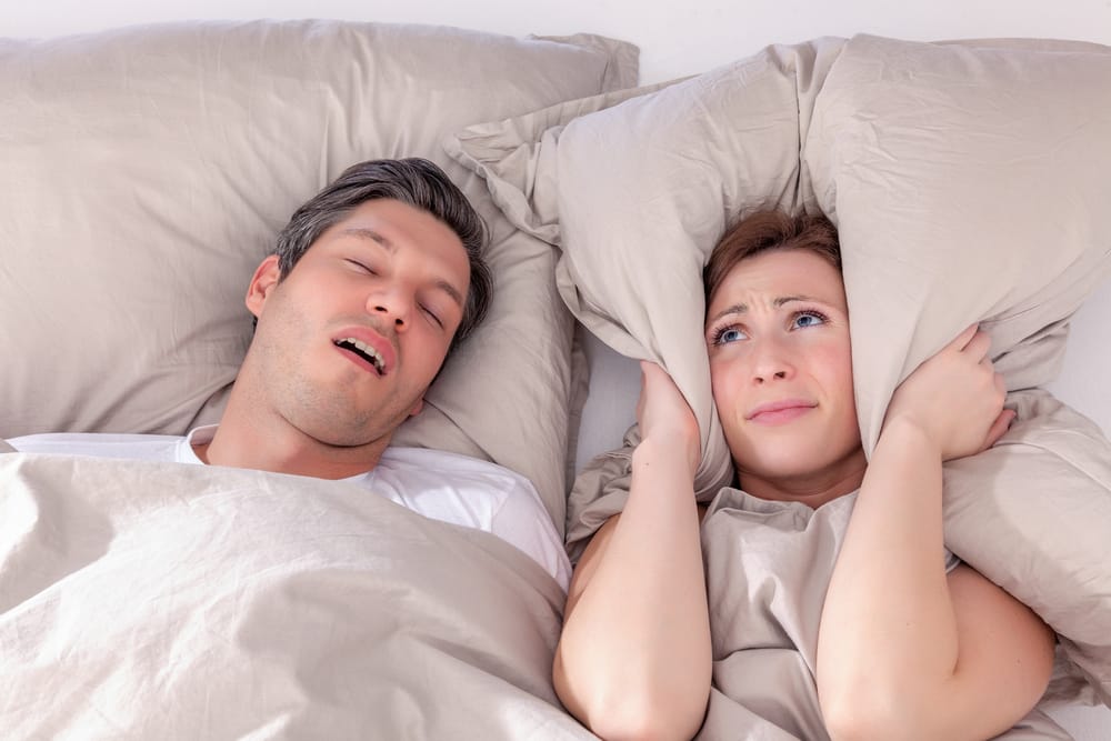 A woman looks frustrated in bed, covering her ears with a pillow while a man beside her snores loudly - Dentist Atlanta